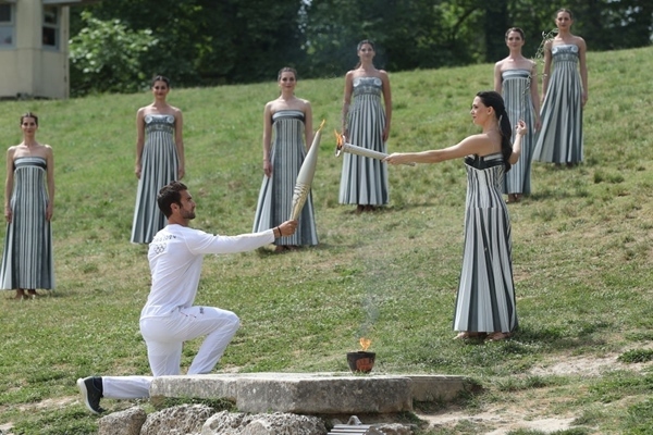 扮演最高女祭司的演員瑪麗·米娜（前右）點(diǎn)燃第一棒火炬手——希臘運(yùn)動(dòng)員、東京奧運(yùn)會(huì)男子賽艇項(xiàng)目金牌得主斯特凡諾斯·恩圖斯科斯手中的火炬。新華社記者 趙丁喆 攝